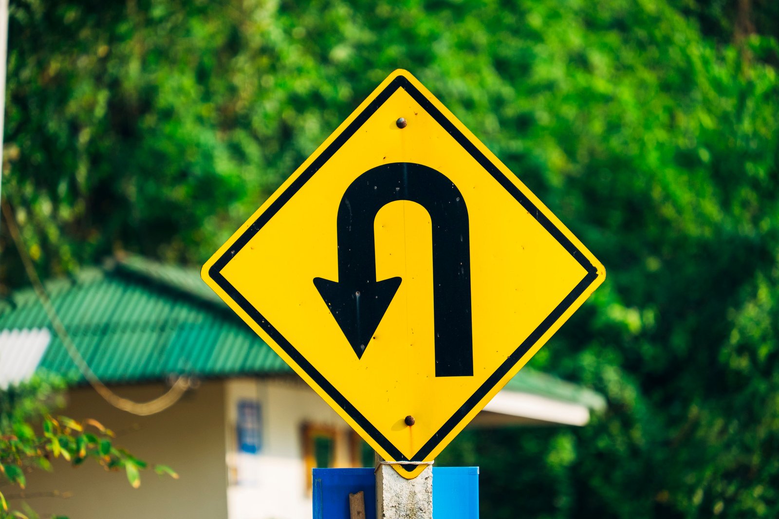 Yellow diamond road sign with a black U-turn arrow, mounted near a green-roofed building amid lush trees, conveying a cautionary tone.