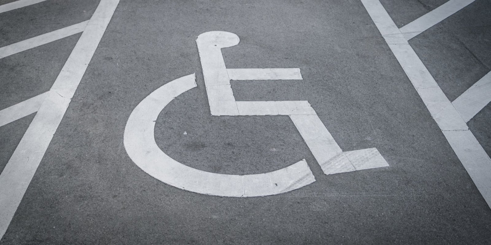 White wheelchair accessibility symbol painted on dark asphalt in a parking lot, viewed from above; high-contrast marking clearly conveys accessible parking.