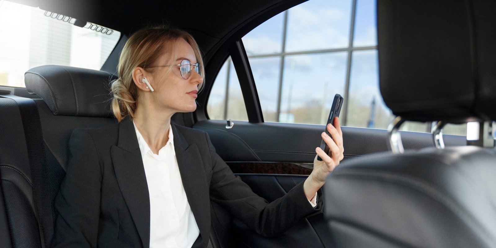 Businesswoman in a black suit and glasses sits in a car at airport, holding a smartphone and gazing out the window with a calm, professional demeanor.