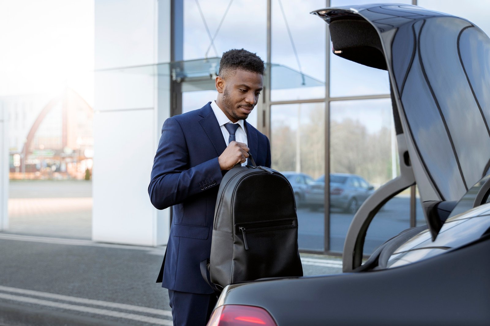 Businessman in a navy suit places a black backpack into the open trunk of a black sedan car outside a modern glass airport; professional travel.