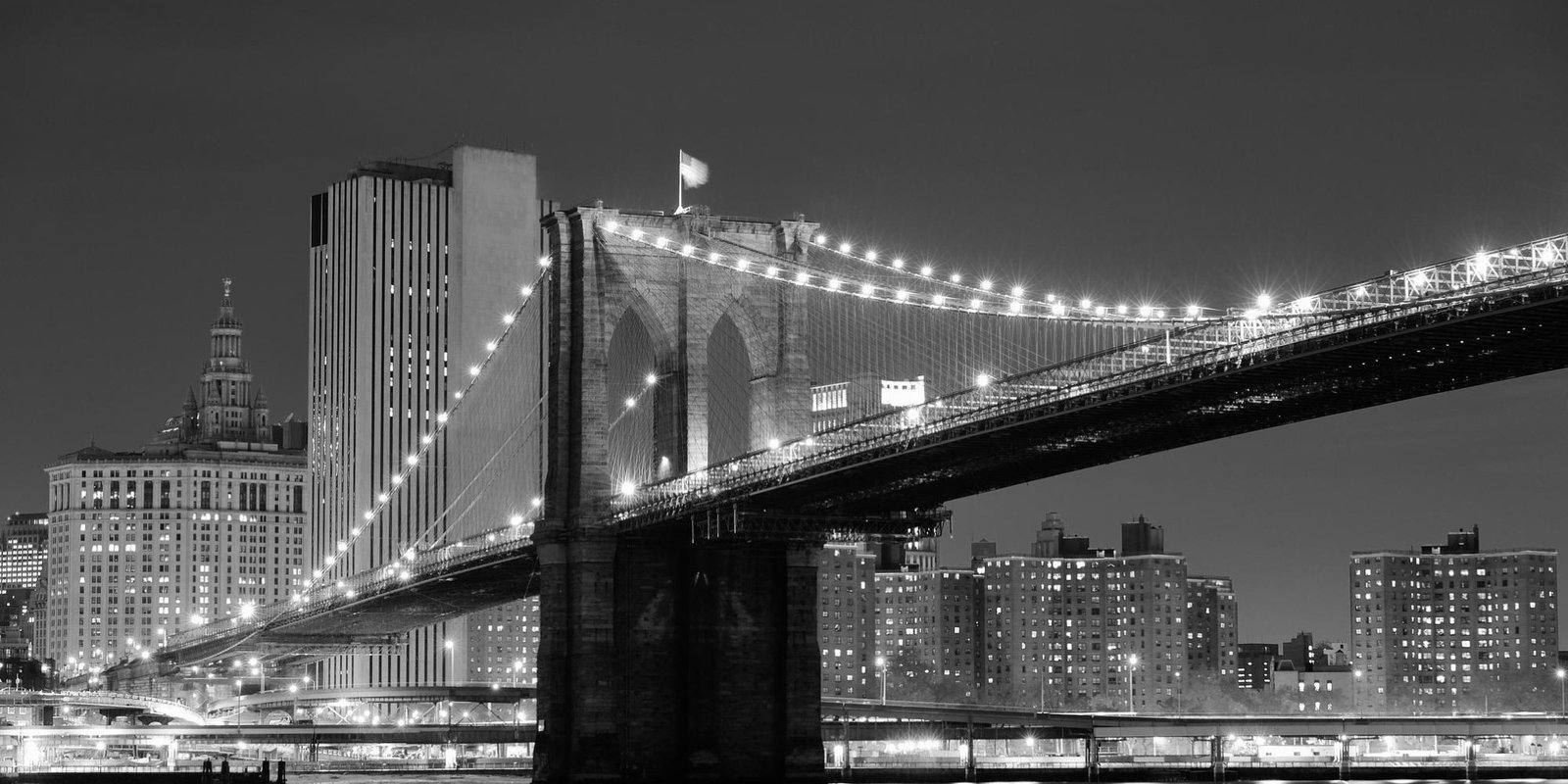 Black-and-white night panorama view of the Brooklyn Bridge lit with strings of lights, spanning the East River beside Manhattan buildings; serene mood.