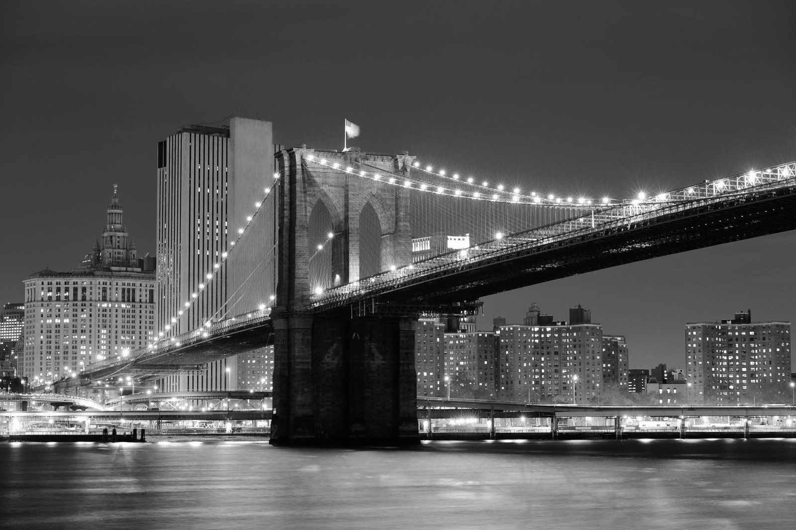 Black-and-white night view of the Brooklyn Bridge lit with strings of lights, spanning the East River beside Manhattan buildings; serene mood.