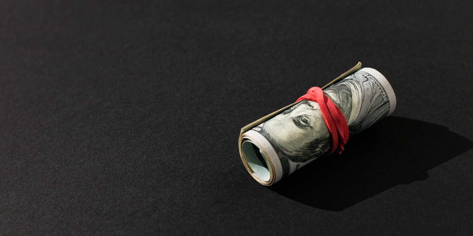 Rolled U.S. dollar bill secured with a red band on a black surface, casting a deep shadow; an intriguing, high-contrast still life.