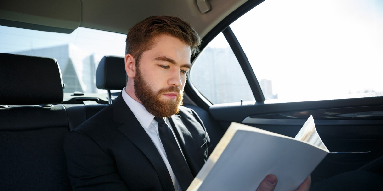 Bearded businessman in a black suit reads a document in the back seat of a car, with a bright city skyline visible through the window.
