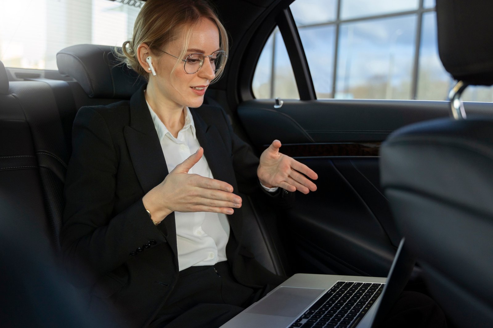 Businesswoman in glasses gestures beside an open laptop in a luxury car, appearing focused and engaged during a professional discussion.