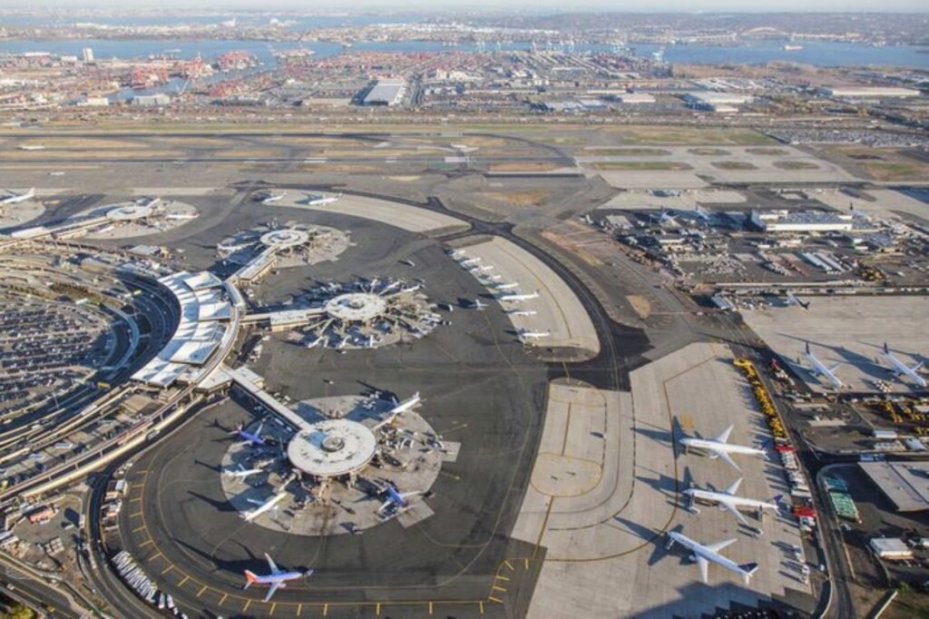 Aerial view of busy coastal Newark Liberty airport, showing multiple terminals, parked passenger planes, runways, taxiways, and a harbor city beyond.