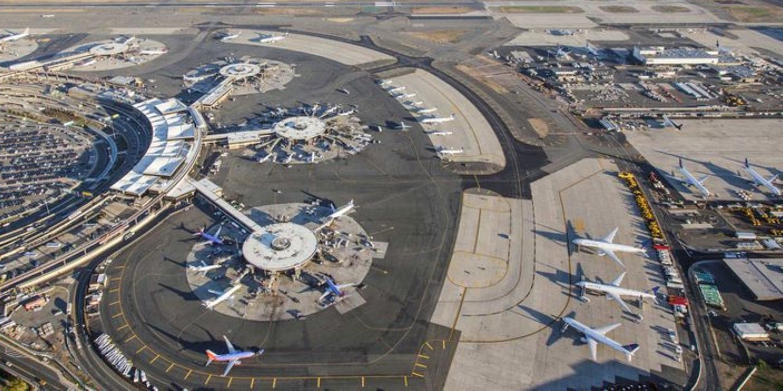 Panorama view of busy coastal Newark Liberty airport, showing multiple terminals, parked passenger planes, runways, taxiways, and a harbor city beyond.