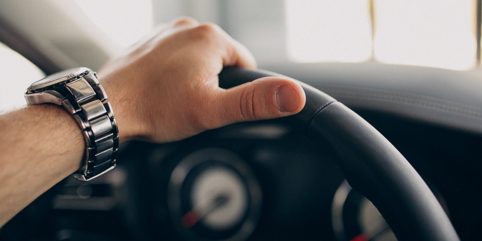Close-up of a driver’s hand wearing a metal wristwatch gripping a car steering wheel, with a blurred speedometer suggesting focus and control.
