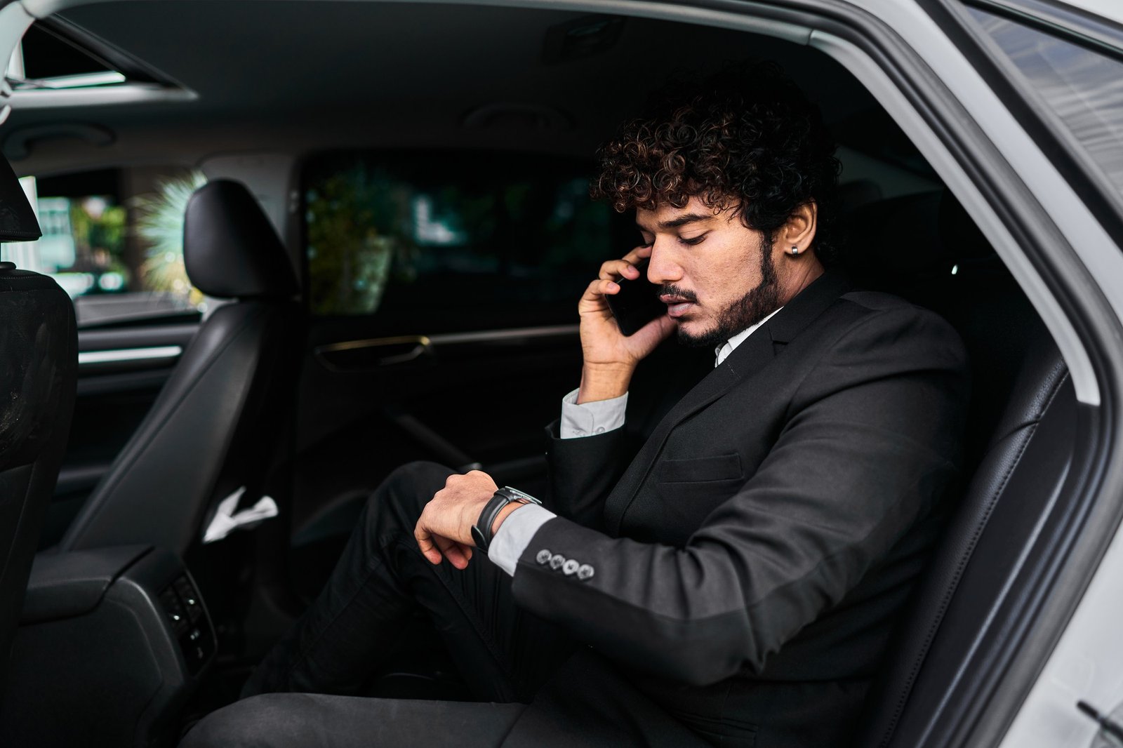 A businessman in a black suit sits in a car’s back seat, speaking on a phone and checking his watch with a serious, thoughtful expression.