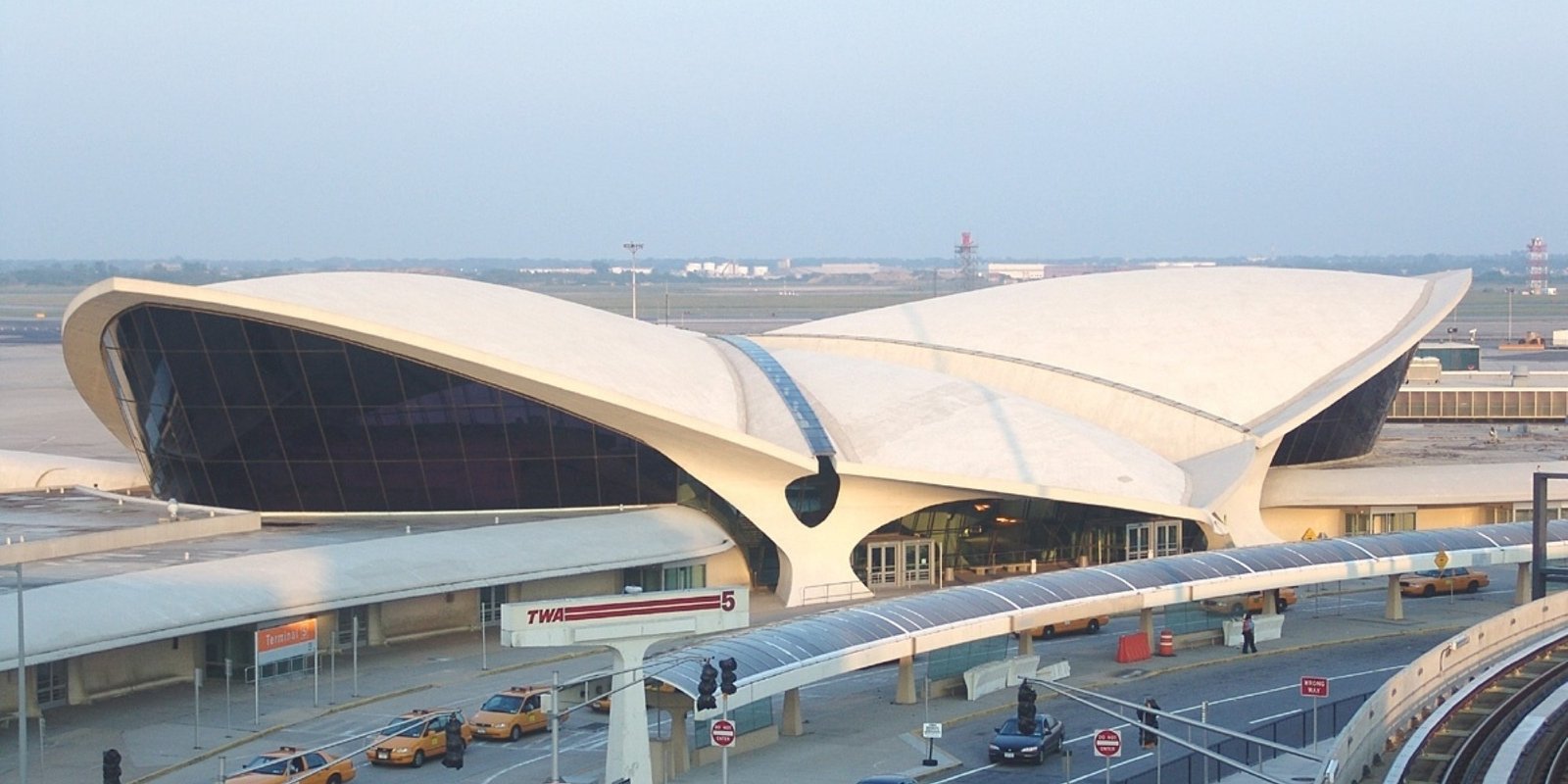 Panorama view of the exterior of John F. Kennedy airport with a dramatic white, wing-shaped roof, glass facade, taxis, and cars along the busy drop-off roadway.
