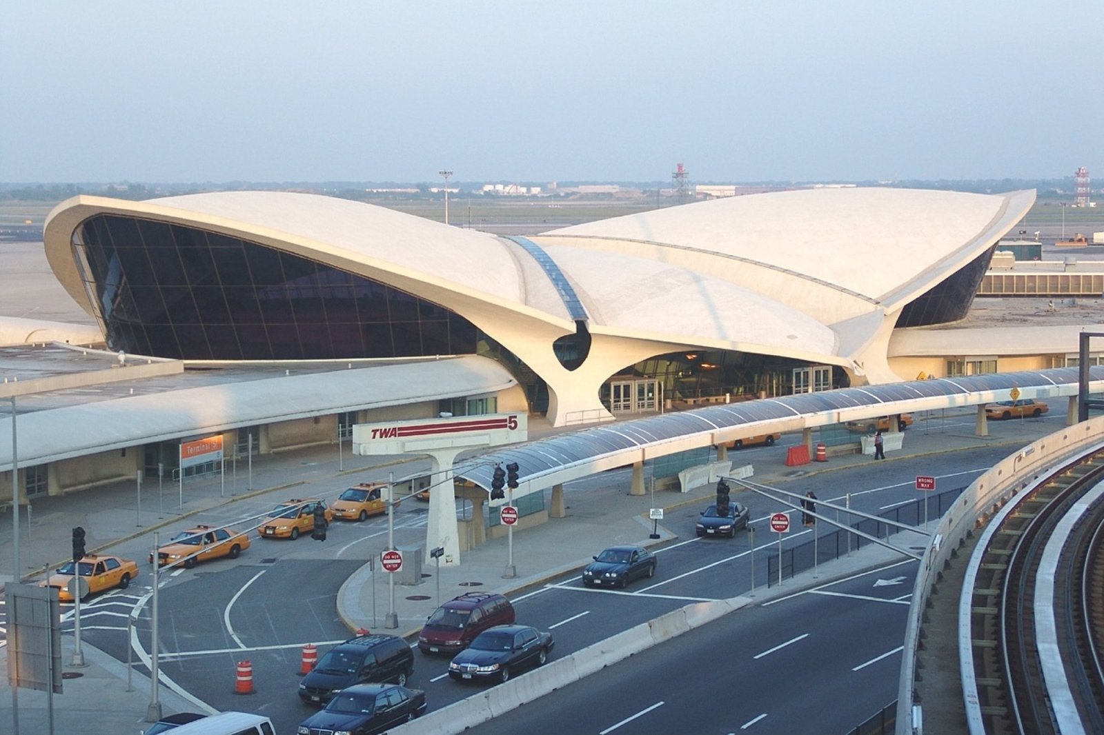 Exterior of John F. Kennedy airport with a dramatic white, wing-shaped roof, glass facade, taxis, and cars along the busy drop-off roadway.