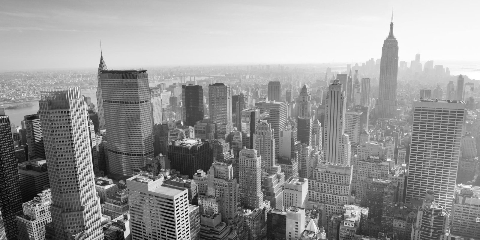 Black-and-white aerial panorama view of Manhattan’s dense skyline, featuring the Empire State Building, towering skyscrapers, and a busy urban cityscape.