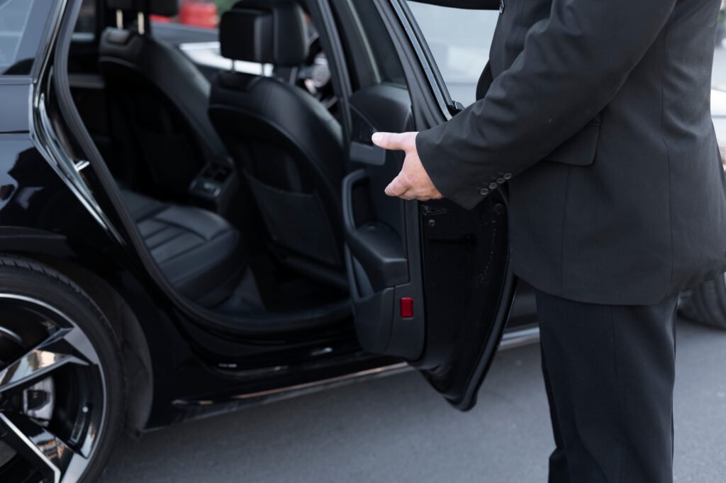 Chauffeur in black suit opens the rear passenger door of a luxury black car, revealing its leather interior; professional chauffeur service.