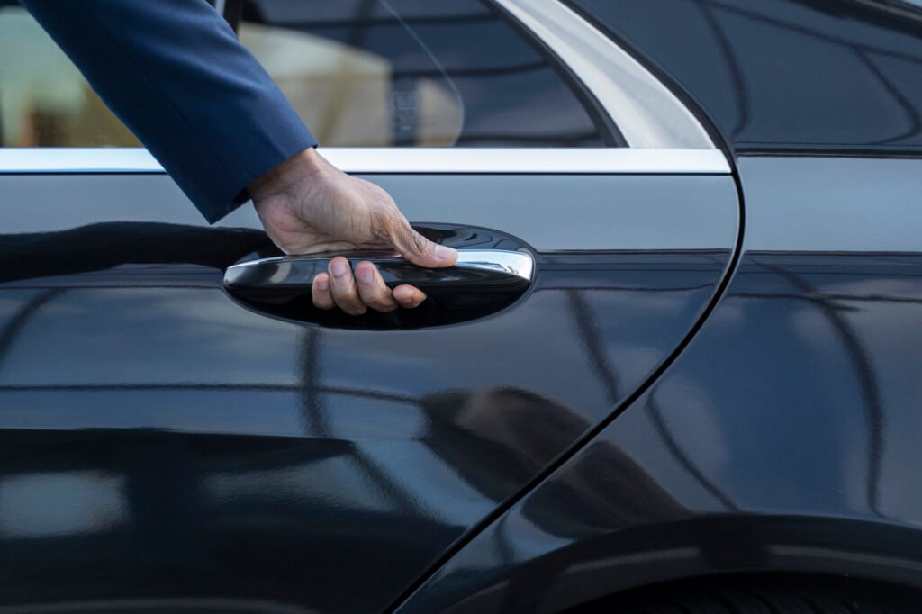 Close-up of a chauffeur's hand in a blue sleeve gripping the chrome handle of a glossy black luxury sedan, ready to open the door; reflections create a sleek tone.