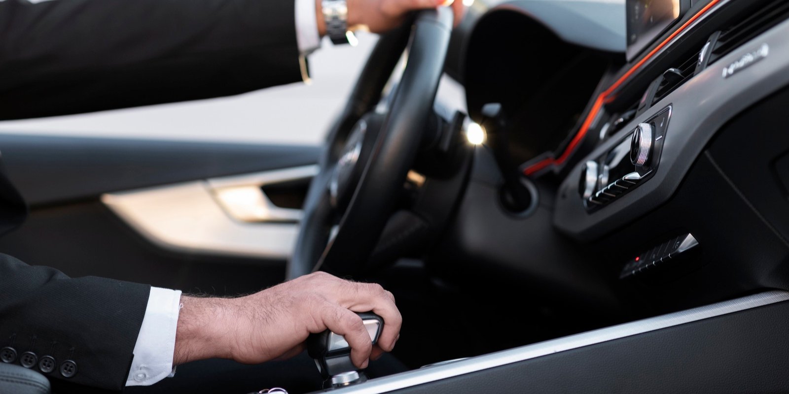 Close-up of a suited driver shifting gears in a sleek luxury car, with one hand on the gear selector and the other on the steering wheel.