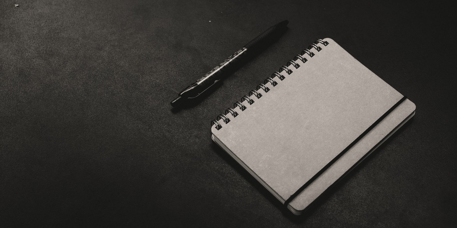 Black-and-white overhead view of a blank spiral notebook and pen on a dark surface, creating a minimalist, thoughtful, professional mood.