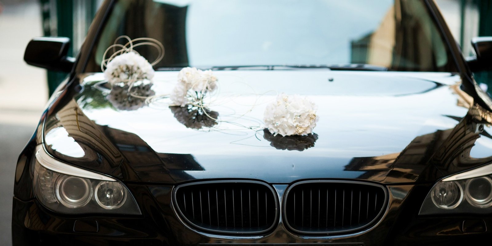 Glossy black luxury sedan with white floral arrangements on its hood, reflecting city buildings; an elegant, romantic wedding atmosphere.