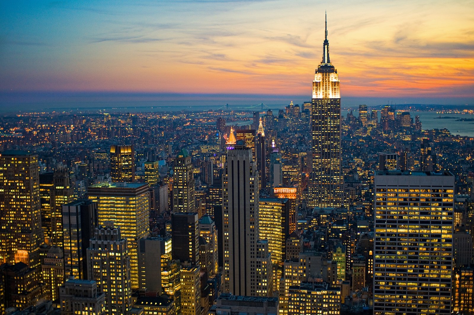 Twilight aerial view of New York, with the illuminated Empire State Building rising above a dense city skyline and glowing streets.