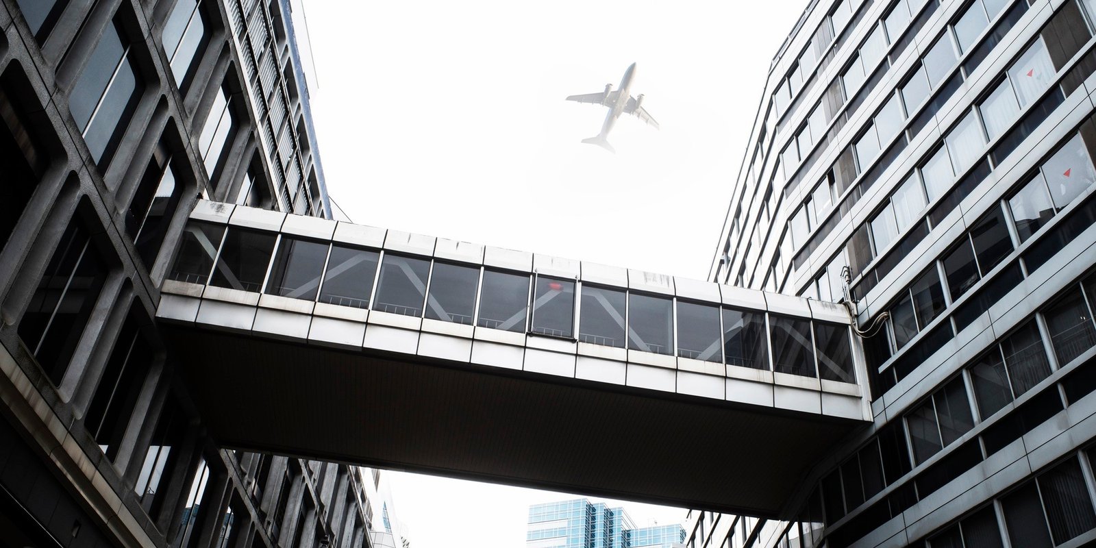 Low-angle view of modern skyscrapers connected by a glass skybridge, with a passenger airplane overhead creating a dramatic urban scene.