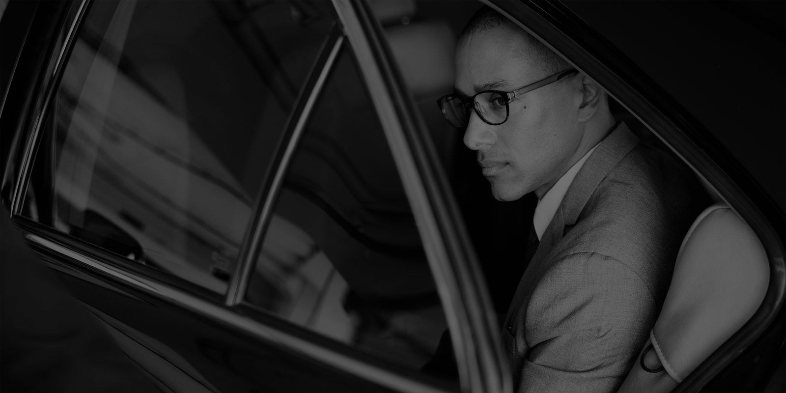 Black-and-white portrait of a businessman in glasses and a suit seated inside a car, viewed through the open rear door, creating a reflective mood.