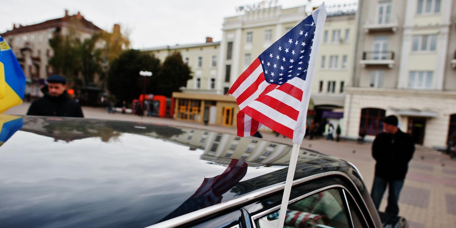 American flag mounted on a car, reflected in its roof, with people and elegant city buildings in a plaza.