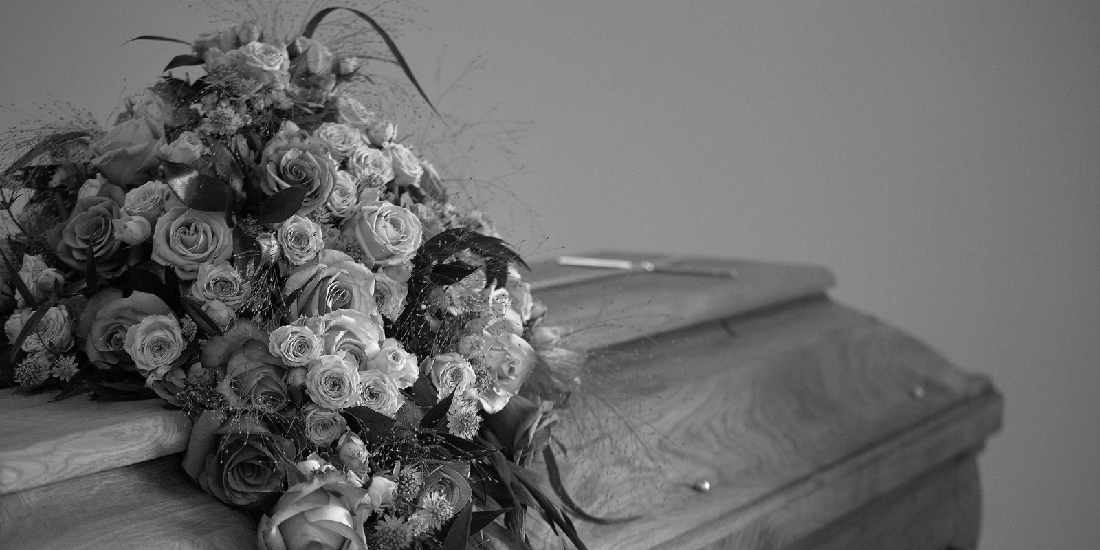 Grayscale close-up of a large bouquet of roses and mixed flowers resting on an ornate wooden coffin, creating a solemn, mournful mood.