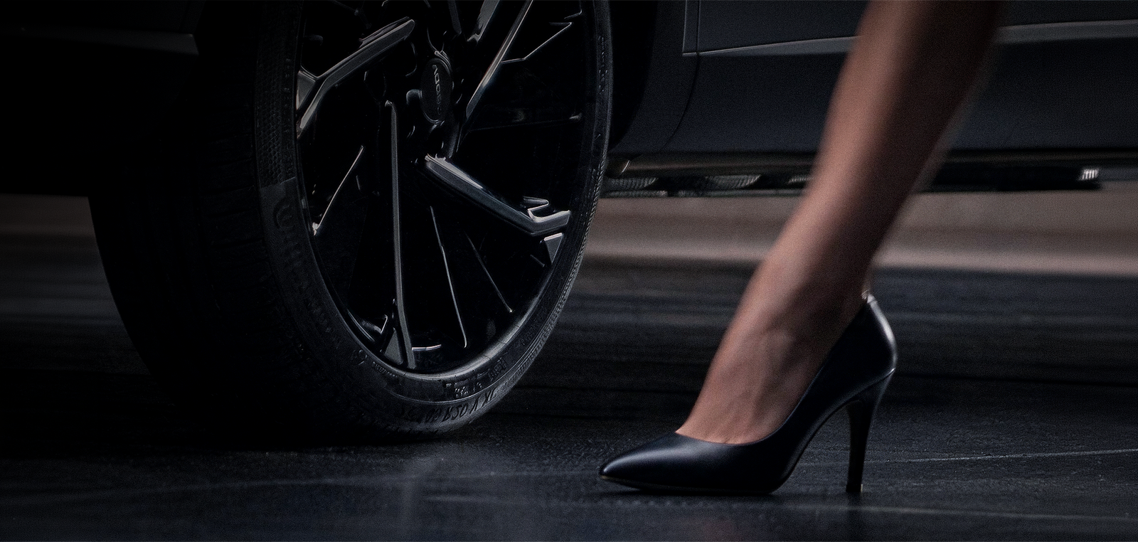 Close-up of a woman’s black high-heeled shoe stepping onto pavement beside a parked car’s wheel, creating a sleek, dramatic mood.