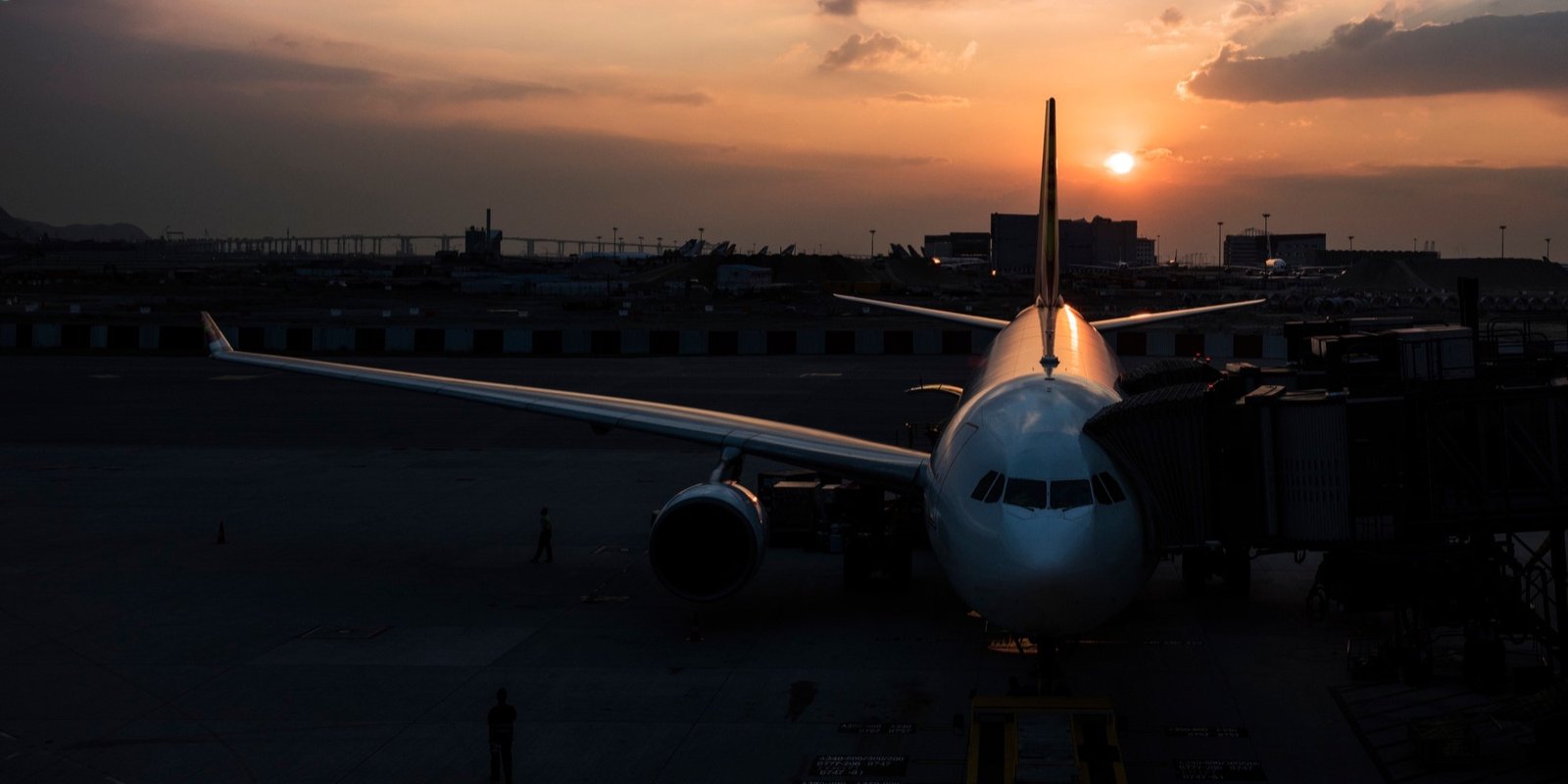 Airliner parked at an airport gate at sunset, with sunlight behind its tail, dark tarmac, terminal structures, and a calm, dramatic mood.