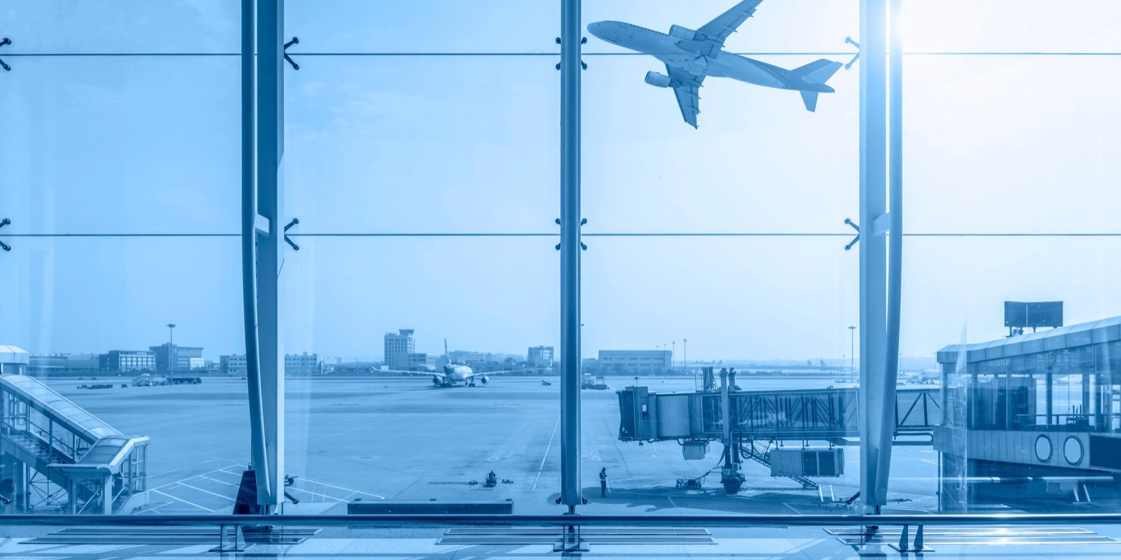 Blue-tinted airport terminal window overlooking a runway, with a passenger airplane flying overhead and a jet bridge in the foreground.