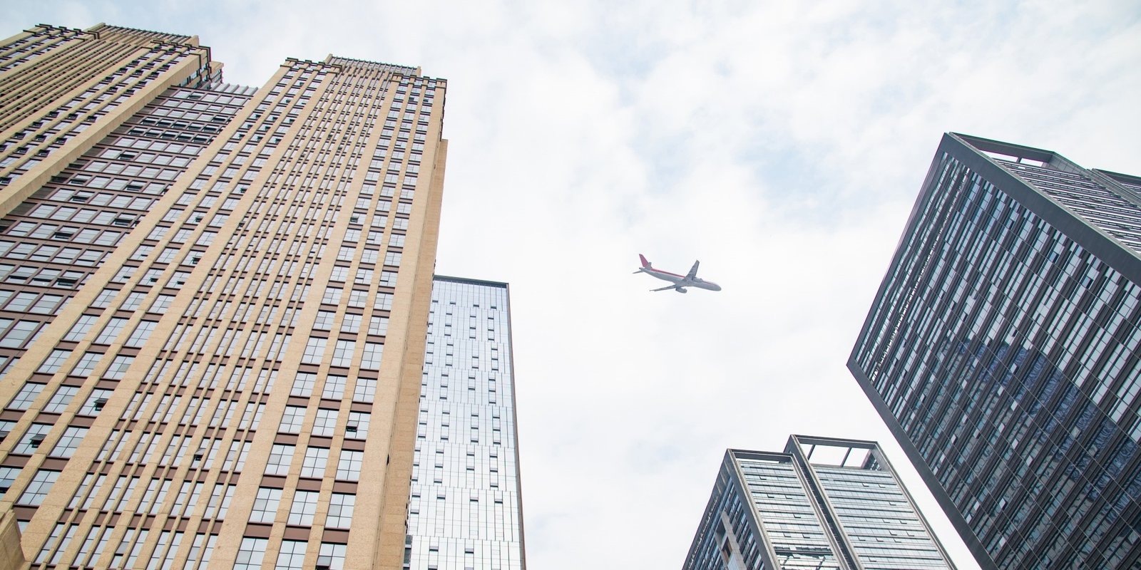 Low-angle view of tall city skyscrapers framing a passenger airplane in a cloudy sky, creating a dramatic urban scene.