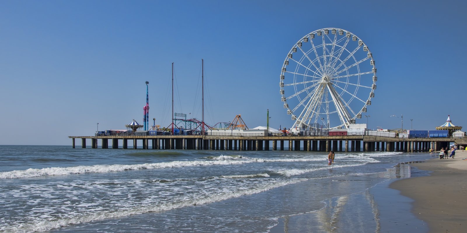 Sunny seaside view of Atlantic City beach with a large Ferris wheel, amusement rides, ocean waves, beachgoers, and reflections on wet sand.