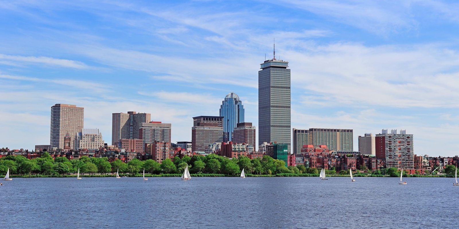 Boston skyline viewed across the Charles River, with sailboats on blue water, a green shoreline, and skyscrapers beneath a bright, partly cloudy sky.