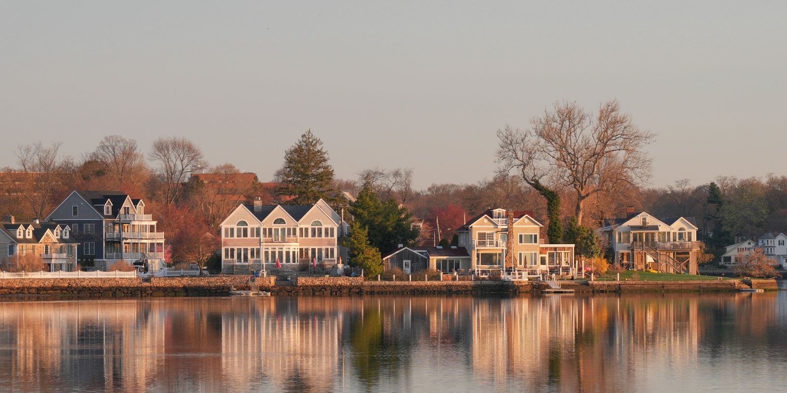 Autumn waterfront Stamford with elegant homes, bare trees, and warm sunlight reflected in the calm lake, creating a peaceful scene.