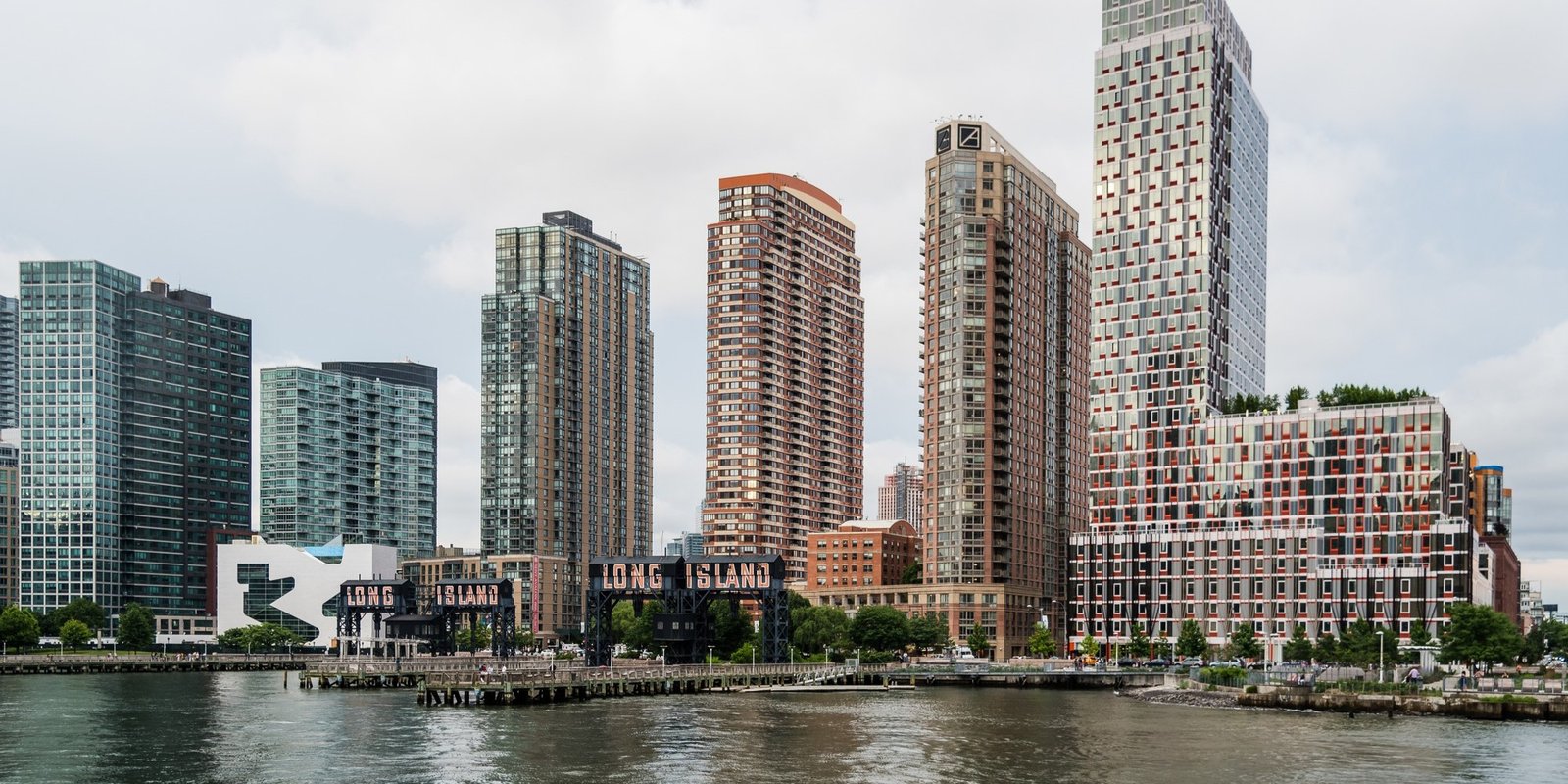 Modern waterfront Long Island skyline with tall glass and brick high-rises, a riverside promenade, and a cloudy sky creating a calm urban mood.