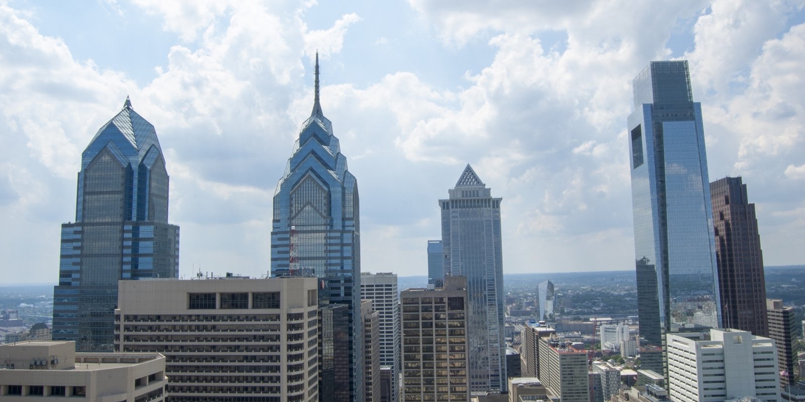 Philadelphia skyline with distinctive glass skyscrapers and dense downtown buildings beneath a bright blue, cloud-filled sky; urban cityscape.