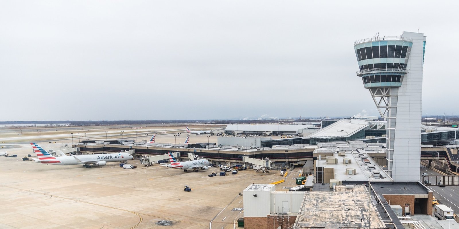 Panoramic overcast airport scene with a tall control tower, terminal gates, parked passenger planes, service vehicles, and a broad winter tarmac.