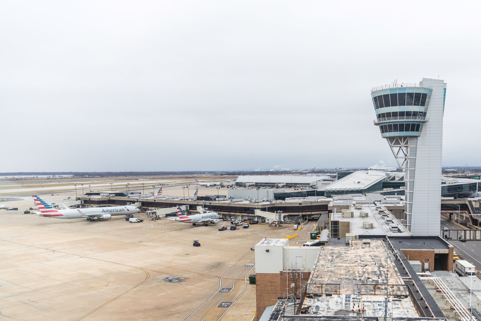 Overcast airport scene with a tall control tower, terminal gates, parked passenger planes, service vehicles, and a broad winter tarmac.