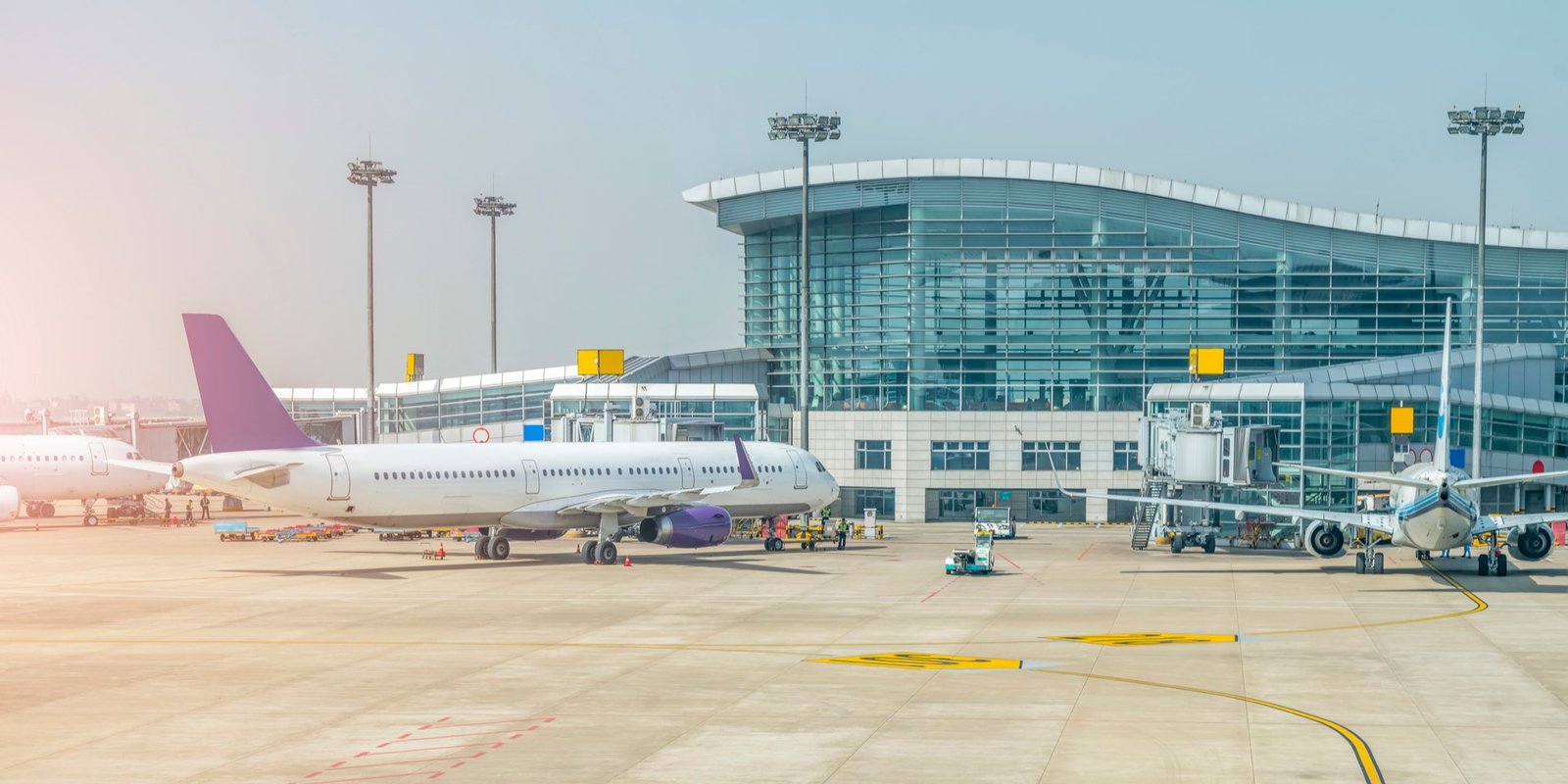 Passenger airplane with a purple tail parked at an airport gate beside a glass terminal, jet bridges, ground vehicles, and blue sky.
