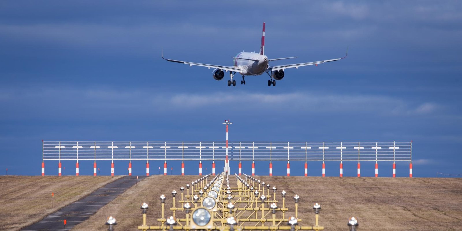 Passenger airplane descending toward a runway, aligned with glowing approach lights across a flat field beneath a dramatic blue, cloudy sky.