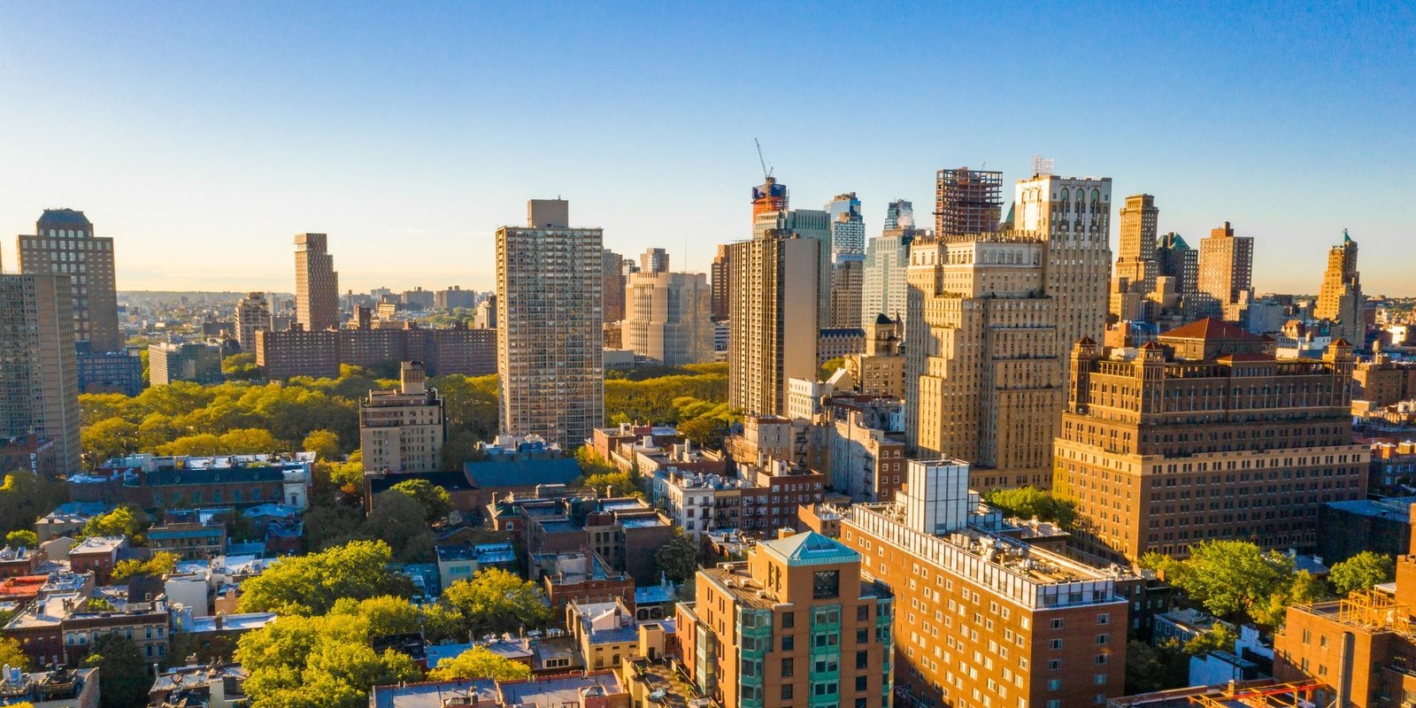 Sunlit city skyline with dense high-rise buildings, rooftops, construction cranes, and leafy neighborhoods beneath a clear blue sky.