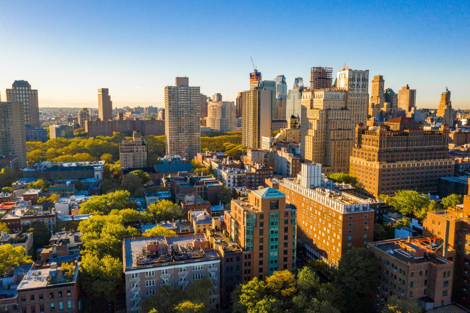 Sunlit city skyline with dense high-rise buildings, rooftops, construction cranes, and leafy neighborhoods beneath a clear blue sky.